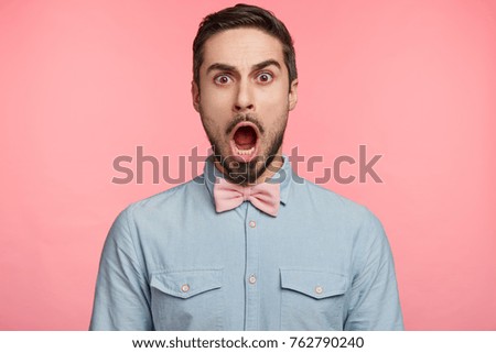 Portrait of attractive brunet male opens mouth widely and looks with bugged eyes, astonished to hear unexpected news, wears pink tie bow, isolated over colorful background. Emotions concept.