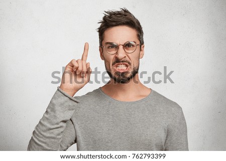 Portrait of annoyed bearded handsome man clenches teeth, frowns face, raises fore finger as proves his rightness and reproaches someone who made big mistake. Irritated furious male enterpreneur
