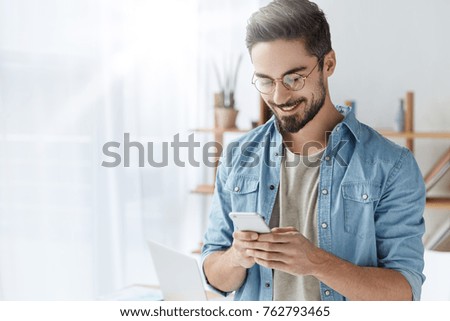 Glad fashionable young bearded guy wears stylish clothes and denim shirt, has trendy hairstyle, happy to exchange messages with friends, uses free internet connection on electronic modern gadget Royalty-Free Stock Photo #762793465