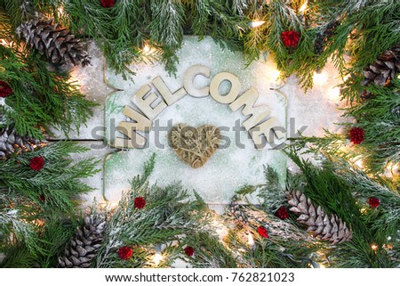 Welcome sign with Christmas tree garland border, string of lights, pine cones, red berries and rope heart on antique rustic wooden snowy background; winter holiday sign with decorations