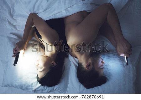 Girl and man are lying on a bed in their underwear and looking at the smartphone. Social networks and loneliness. A girl with a guy on a bed in a dark room with a phone in hand.
