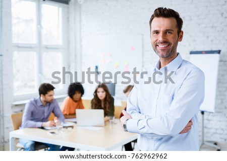 Portrait of happy teacher standing in classroom with students in background Royalty-Free Stock Photo #762962962