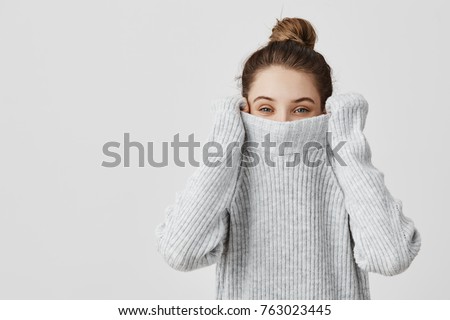 Portrait of girl pulling her trendy sweater over head having fun. Woman with tied hair in topknot being childish disappearing in her clothes looking from underneath. Happiness concept Royalty-Free Stock Photo #763023445