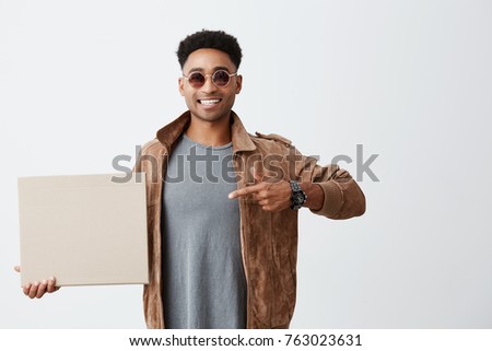 Positive emotions. Close up of young good-looking afro american male student with curly hair in casual clothes and sun-glasses holding carton board, pointing at it with hand, smiling with teeth.