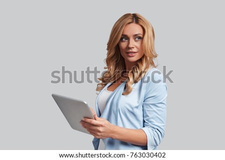 Searching for fresh ideas. Attractive young woman using digital tablet and smiling while standing against grey background