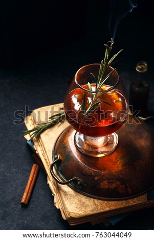 Whiskey cocktail with ice and burnt rosemary. Dark background, vintage papers, dramatic ligting. Vertical composition.