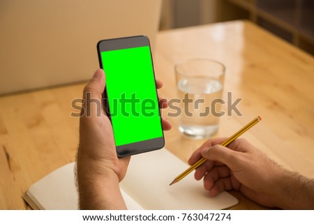 Young adult man sitting at wooden desk writing on notebook with pencil hold green screen smartphone with glass of water in bright office in the morning.