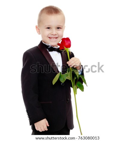 Beautiful little boy in a strict black suit , white shirt and tie.Boy holding a flower of a red rose on a long stem.Isolated on white background.