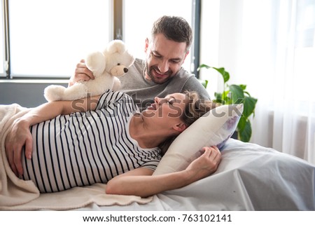 Good morning, my darling. Portrait of excited man awaking his wife while giving teddy bear to her. Pregnant sleepy woman is lying on bed and smiling