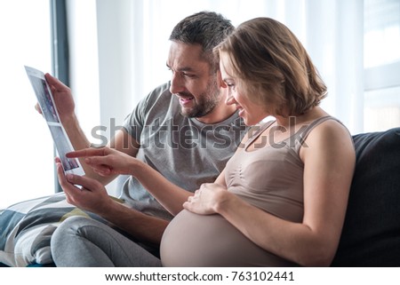 Side view of cheerful family observing ultrasound photo of their baby with interest. Pregnant woman is touching her big belly and smiling