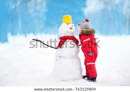Little boy tell snowman about his secrets or gifts for Christmas. Active outdoors leisure with children in winter. Kid during stroll in a snowy winter park