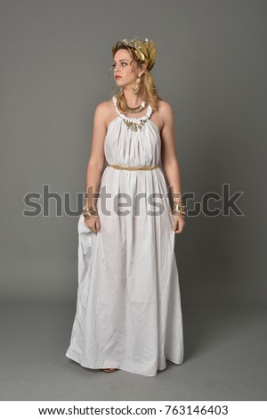 full length portrait of girl wearing white ancient Greek or Roman costume, standing pose on a grey background.
