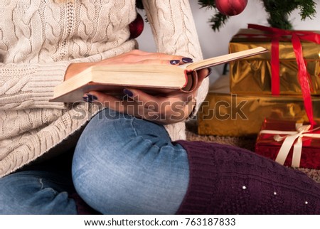 Girl in sweater holding book in hands and Christmas tree and gift box in background, close up, selective focus