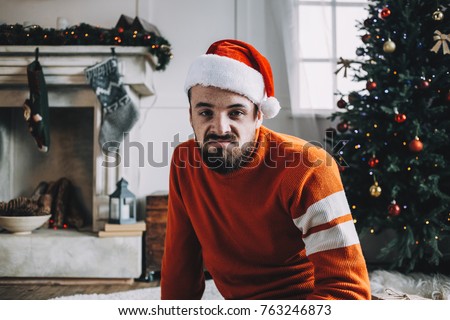 Funny picture of a bearded guy sitting on the floor in a white decorated room. Somehow he is not happy with something because his face shows such emotions as boring and unhappiness. He is looking