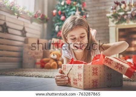 Happy holidays! Cute little child opening present near Christmas tree. The girl laughing and enjoying the gift. 