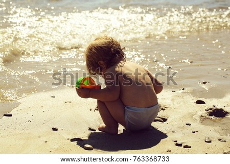 Little curious smart blonde baby boy in sitting on sand sunny day outdoor playing with plastic toys on natural wavy ocean water beach background, horizontal picture