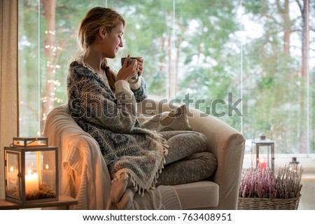 Young beautiful woman sitting home in the chair by the window with cup of hot coffee wearing knitted warm sweater. Cozy room decorated with lanterns and candles. Stay home Royalty-Free Stock Photo #763408951