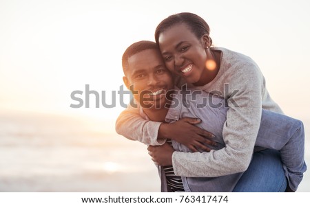 Young African man carrying his smiling girlfriend on his back while enjoying a late afternoon together at the beach Royalty-Free Stock Photo #763417474