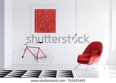 Lamp above red armchair in bright interior with black and white carpet and red painting on the wall above bike