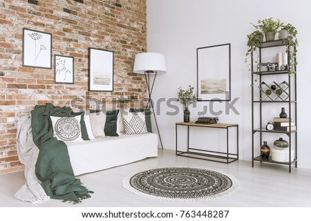 Cushions and blankets on white sofa in cozy living room with pictures on brick wall