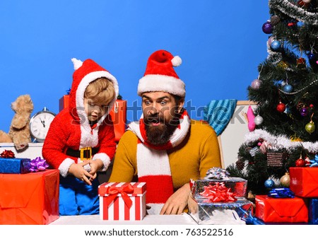Man with beard and surprised face plays with son. Family holidays concept. Christmas family opens presents on blue background. Santa and little assistant among gift boxes near Christmas tree