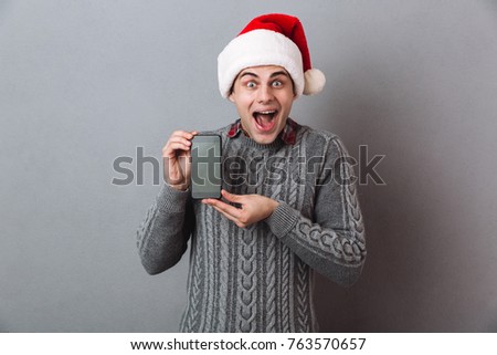 Picture of young excited screaming emotional man wearing christmas santa hat standing isolated over grey wall. Looking camera showing display of mobile phone.