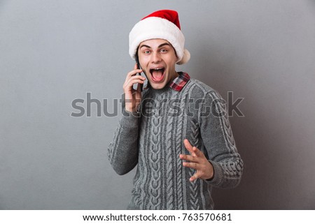 Image of young happy emotional man wearing christmas santa hat standing isolated over grey wall. Looking camera talking by phone.