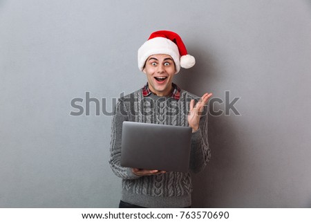 Picture of young emotional man wearing christmas santa hat standing isolated over grey wall. Looking camera using laptop computer.