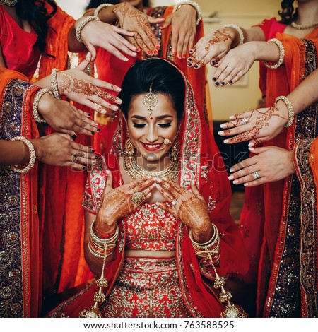 Stunning Indian bride dressed in Hindu traditional wedding clothes lehenga embroidered with gold and a veil sits on the chair while bridesmaids hold their hands with henna tattoos around her Royalty-Free Stock Photo #763588525