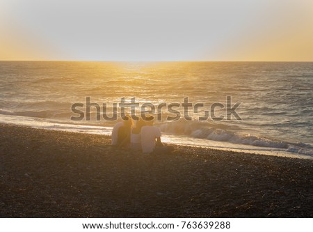 Rear view of a couple silhouette sitting cuddling and enjoying pointing at sun at sunset outside on the beach in winter