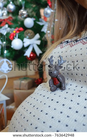 Pregnancy, winter holidays and people concept - pregnant woman looks at the reindeer on her stomach. Christmas tree background