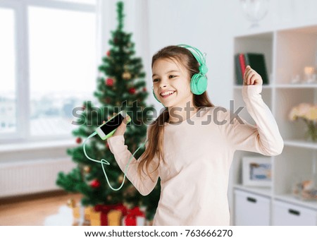 holidays, children and technology concept - happy smiling girl in headphones with smartphone and listening to music and dancing over christmas tree background