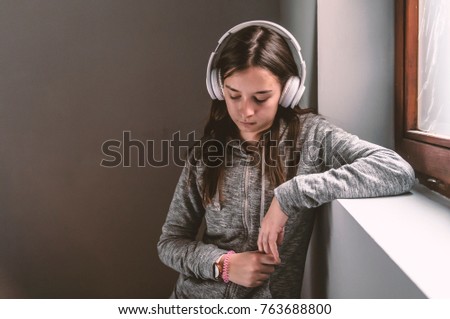 Sport girl Wearing Stereo Headset to Listening Music and Looking Outside. People, technology and teens concept - sad teenage girl sitting on windowsill with earphones listening to music. 