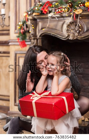 Mother kissing her cute little girl with Christmas gift near a fireplace with decorations