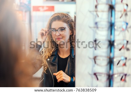 Smiling young woman trying on glasses on mirror in optician. Royalty-Free Stock Photo #763838509