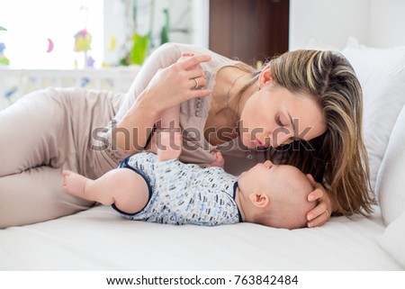 Young mother, lying in bed with her newborn baby boy, playing and interacting together. Family happiness concept. Back light fron the window behind