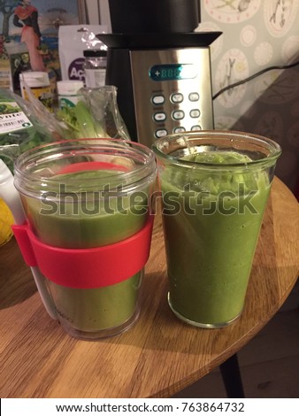 Portrait picture of two green smoothies in plastic cups with a pink band on a table