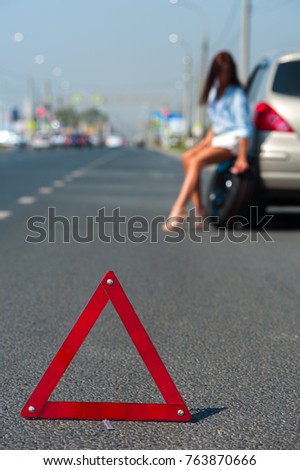 Red triangle - emergency stop sign on the background of blurry woman - driver sits on discarded wheel of her car and waiting for help on the highway on warm summer day.