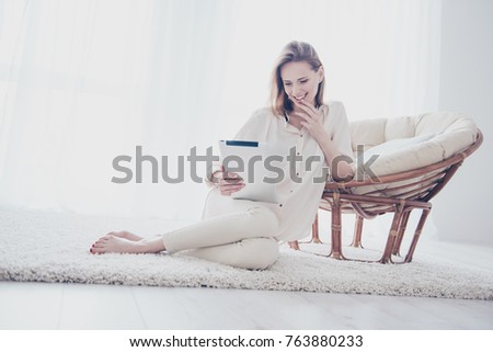Cute lovely beautiful charming attractive woman is sitting on the floor, leaning on the armchair, holding digital tablet and choosing brand new furniture for her house