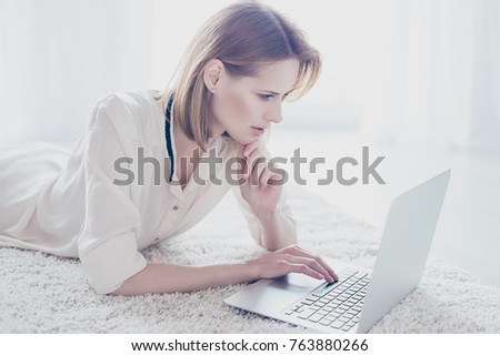 Close up photo of pretty confident pensive young woman with short blonde hair which is lying on the carpet at home and using the laptop for working and ordering goods