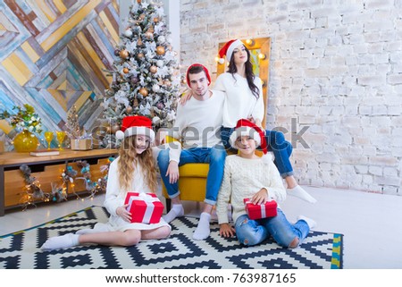 A Christmas photograph of a beautiful family with two children in Santa Claus red caps and red presents. Sit on the couch in the Christmas interior.
