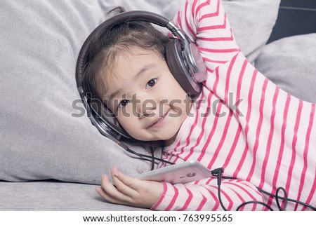 The lovely little girl lying on the sofa and listening to the music is smiling