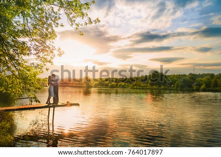 Beautiful summer picture on the nature by the river. A loving couple is standing on the pier on a sunset background.