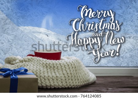 A beautiful image of a red cup, a white scarf and a gift on a window sill with a beautiful winter landscape outside the window. Added text.