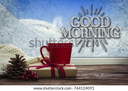 A beautiful image of a red cup, a white scarf and a gift on a window sill with a beautiful winter landscape outside the window. Added text Good morning.