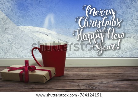 A beautiful image of a red cup, a white scarf and a gift on a window sill with a beautiful winter landscape outside the window. Added greeting text.