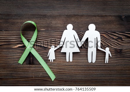 Green ribbon for Lyme disease, kidney cancer, organ donation awareness near paper silhouette of family on dark wooden background top view