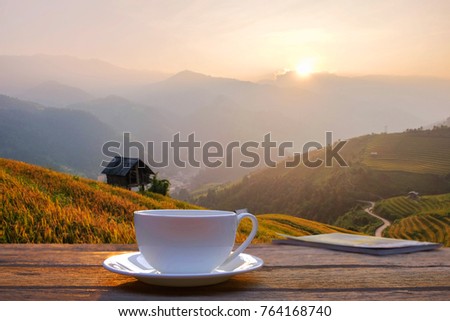 a selective focus picture of a cup of coffee on wooden fence beside terraced rice field