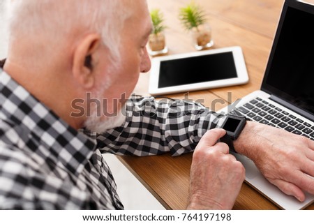 Senior man using laptop, tablet and modern watch with blank screen side view, mockup. Mature male surfing net with computer at home, watching the time, copy space
