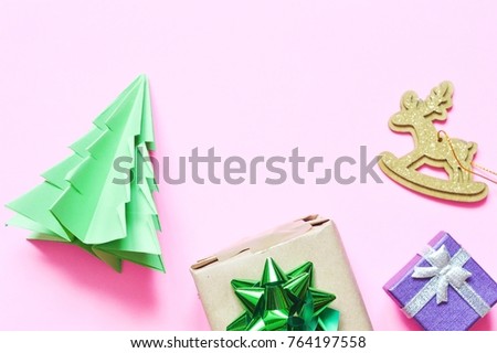 A small Christmas tree made of green paper made in origami style, a gift wrapped in craft paper with a glossy iridescent green bow, a purple gift box and a toy deer on a pink background. Flat lay